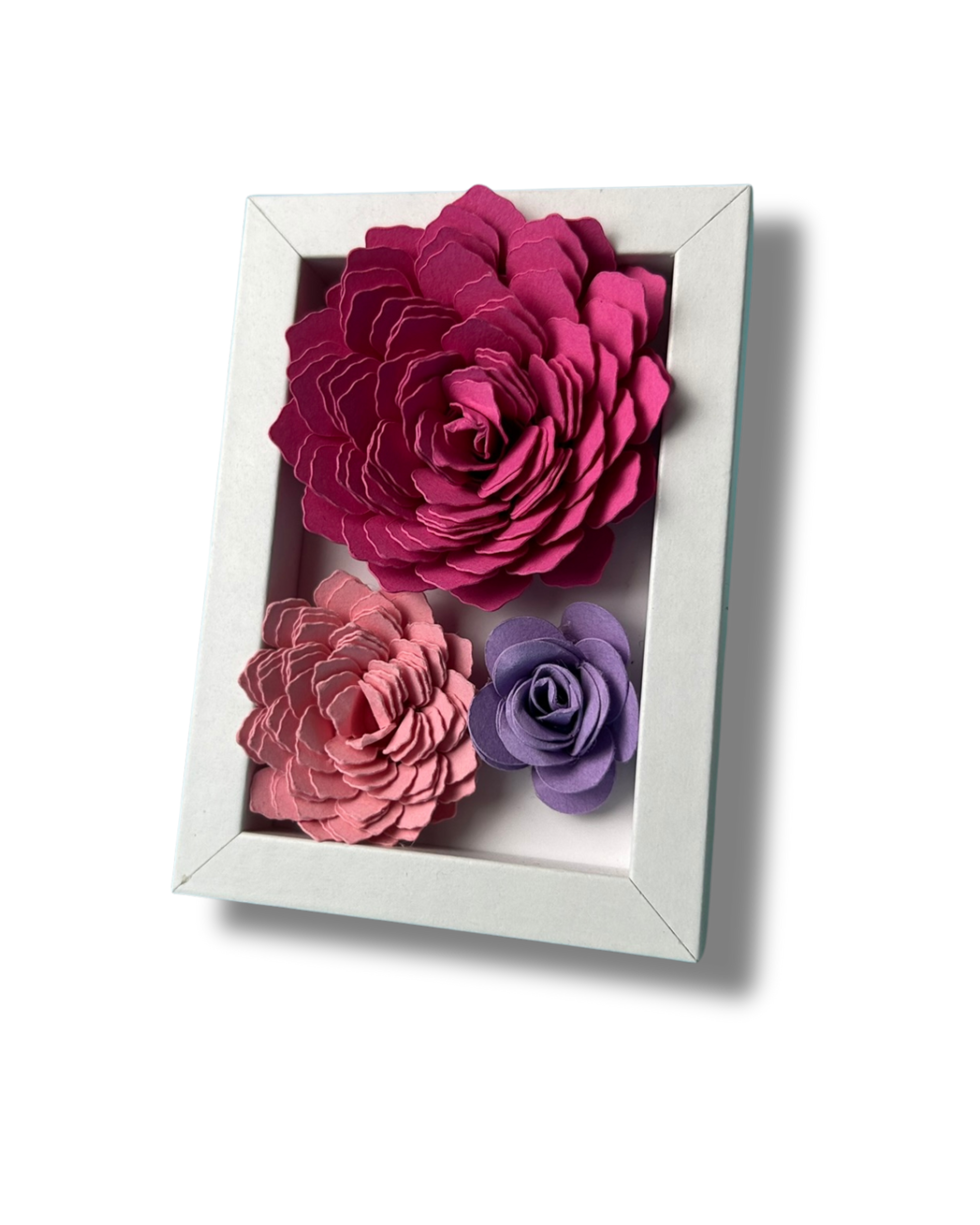 Kleiner weißer Bilderrahmen mit drei Blumen in Pink,lila und rosa vor weißem Hintergrund.