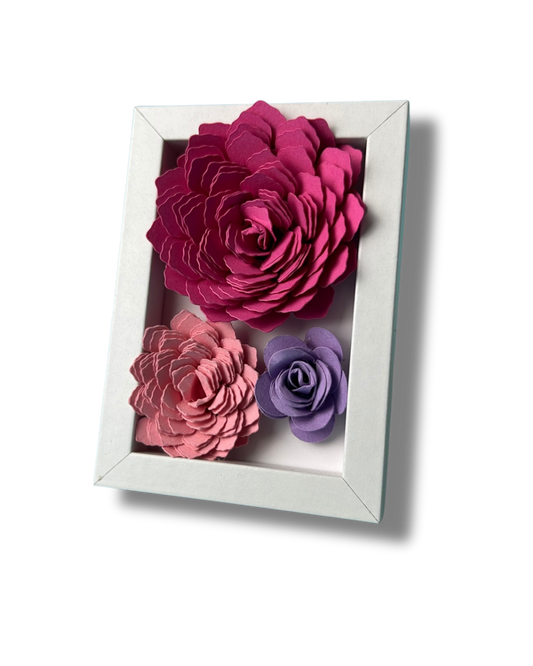 Kleiner weißer Bilderrahmen mit drei Blumen in Pink,lila und rosa vor weißem Hintergrund.