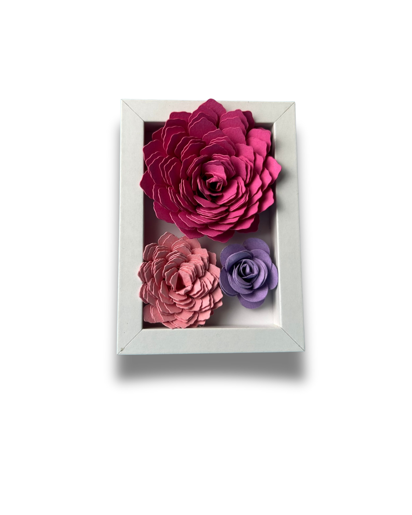 Kleiner weißer Bilderrahmen mit drei Blumen in Pink,lila und rosa vor weißem Hintergrund.